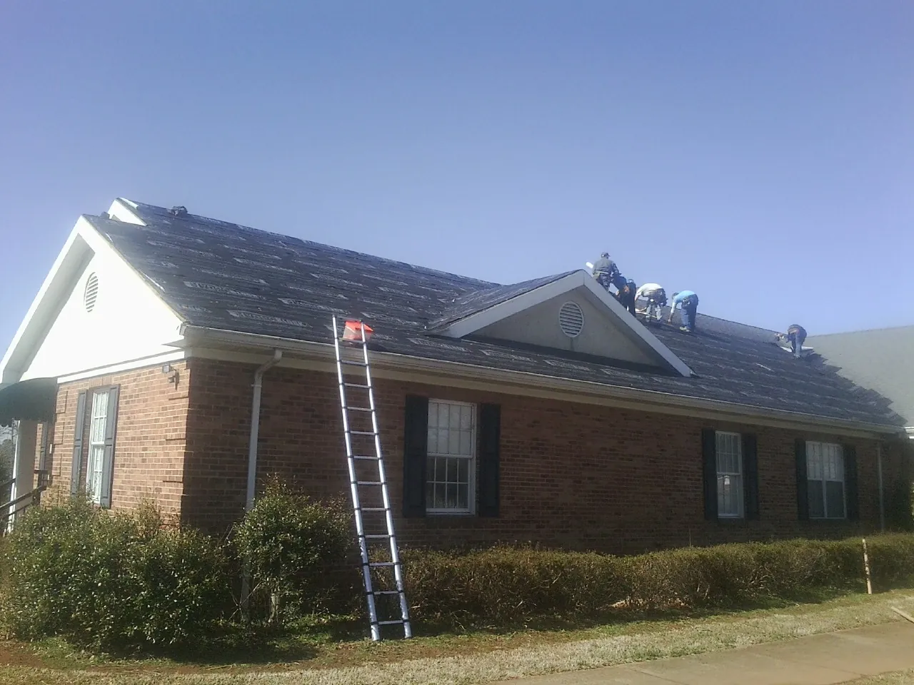 Crew tearing shingles off a brick ranch roof with a ladder set — Emergency Roof Repair work by our crew in East Schodack