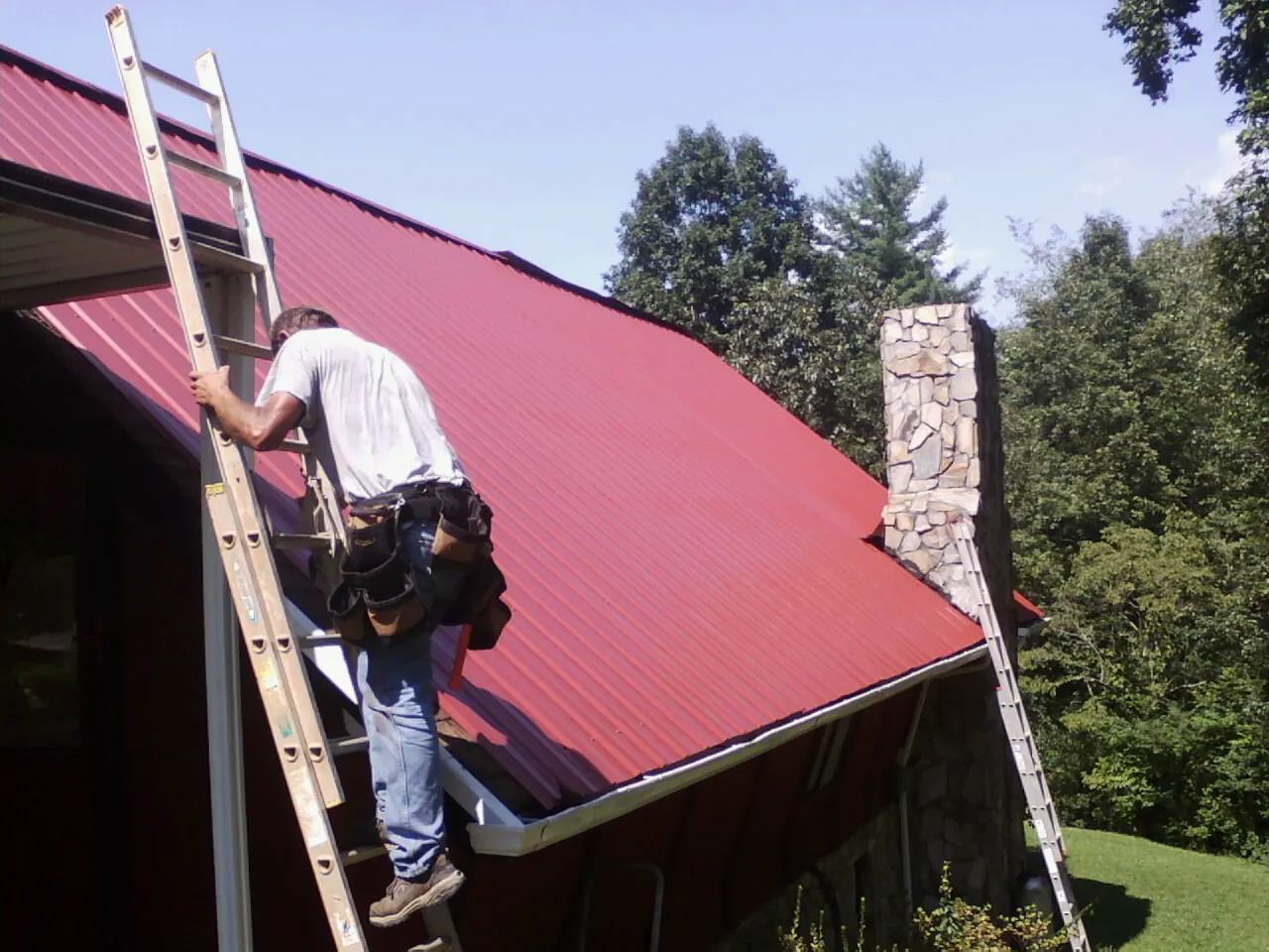 Roofer on a ladder beside a red metal roof and stone chimney — Soffit & Fascia Repair by Roofing Brothers in East Schodack